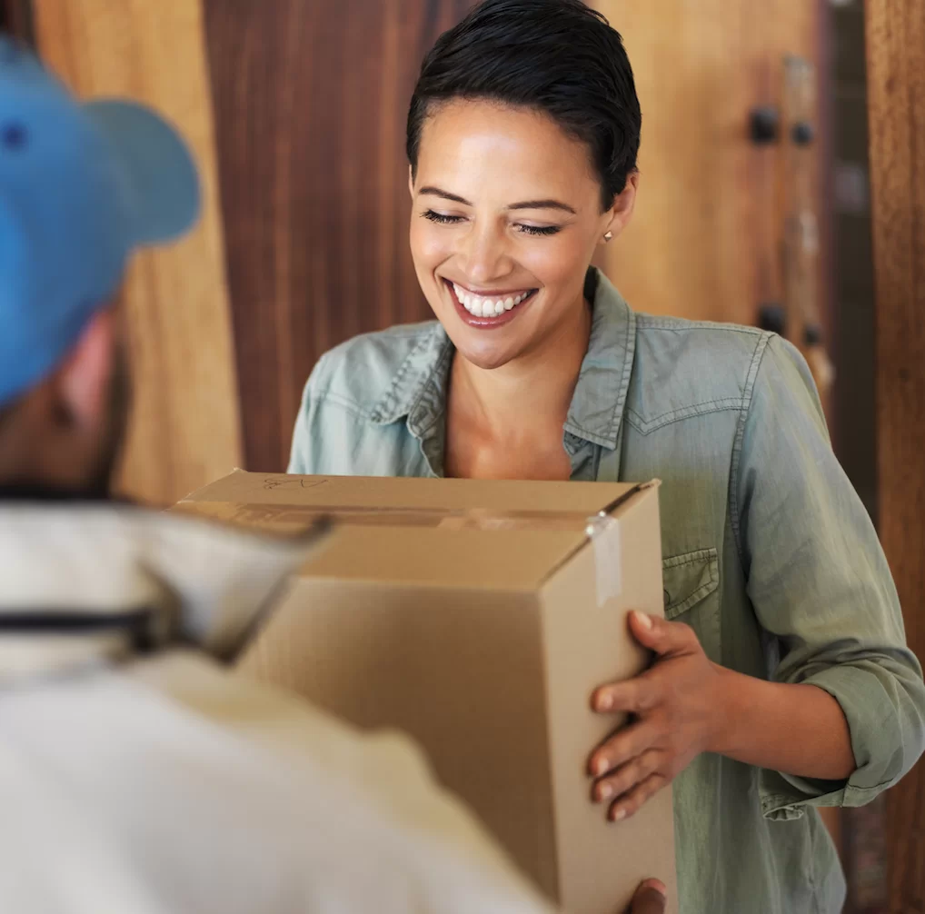 A woman of mixed descent smiling and being handed a brown cardboard box by a delivery person in a blue cap