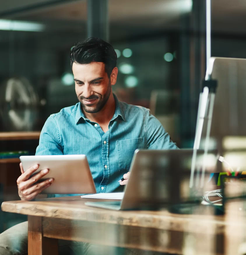 A white man in a denim shirt sits with his tablet and a laptop open at a desk, with a tablet pen, smiling at what he is noting down