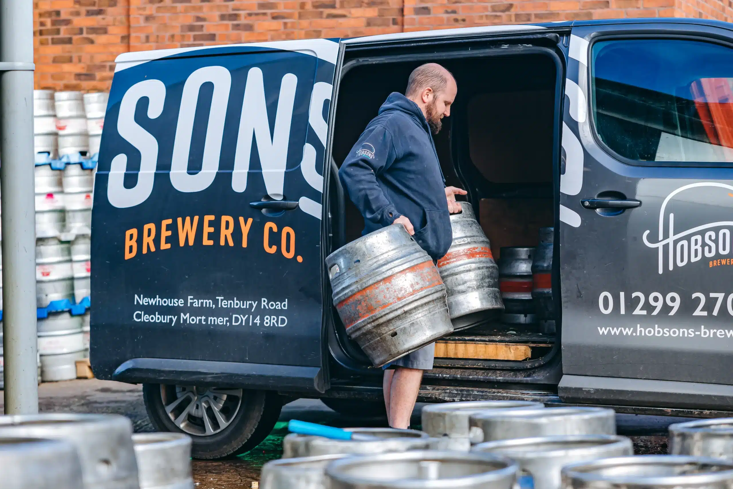 A man loads two kegs of beer into a blue van with Hobsons Brewery branding on the side