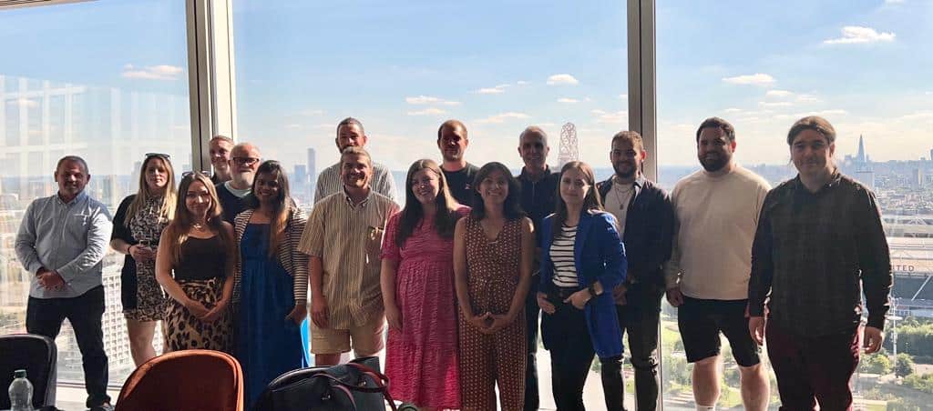 Two rows of people pose for a photograph in front of windows overlooking a cityscape.