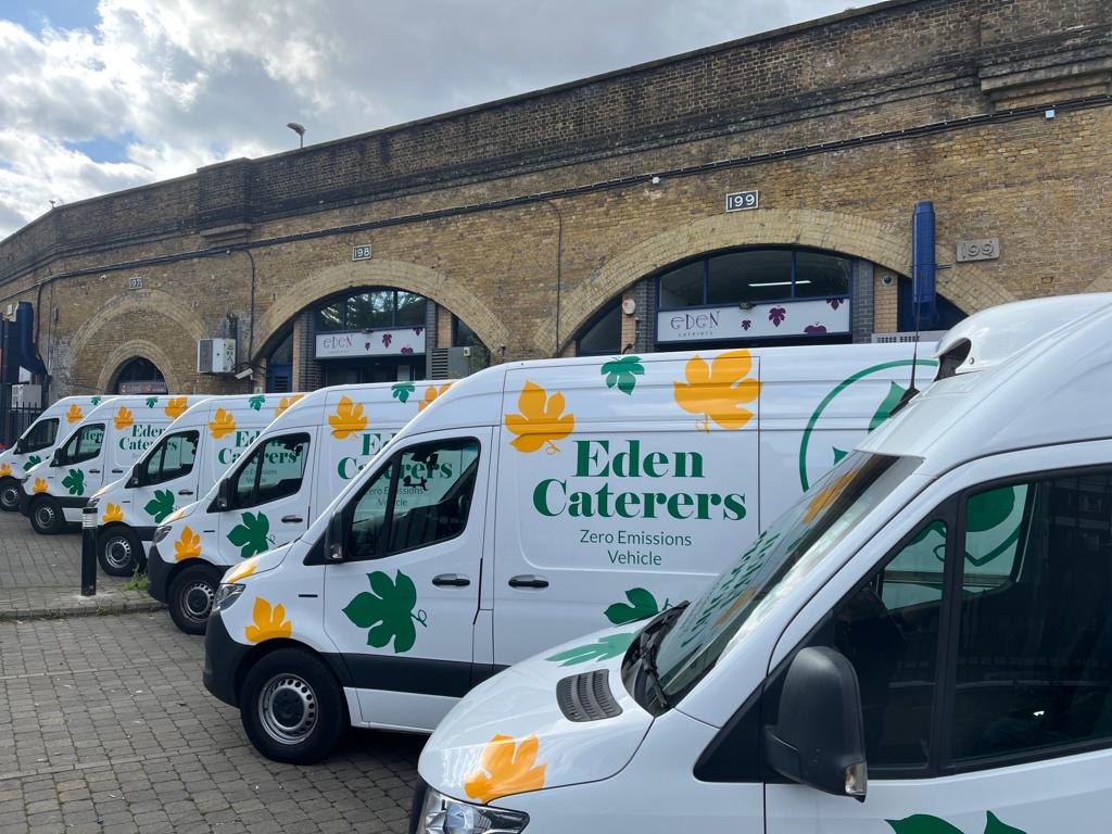 Image displaying Eden Caterers fleet of delivery vans, displaying their logo and green and yellow brand leaves. With a backdrop of their bricked warehouses.