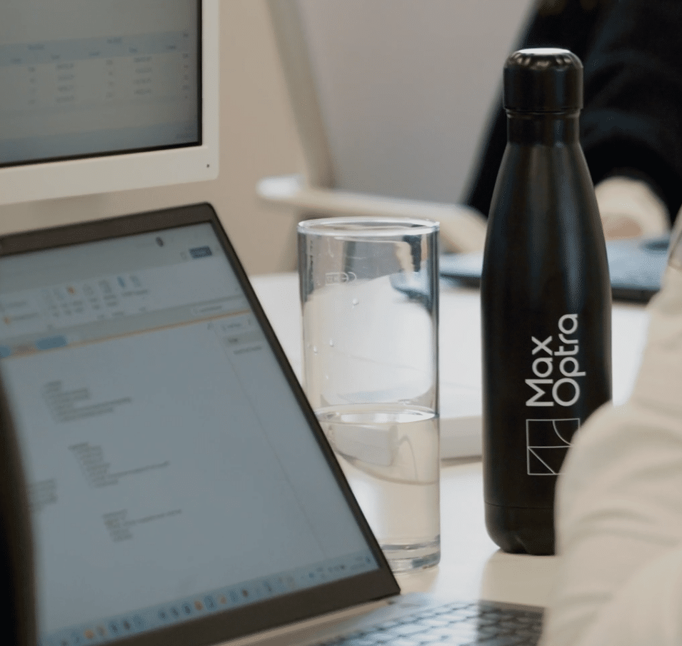 A black MaxOptra branded drink bottle next to a glass of water and a laptop on a desk
