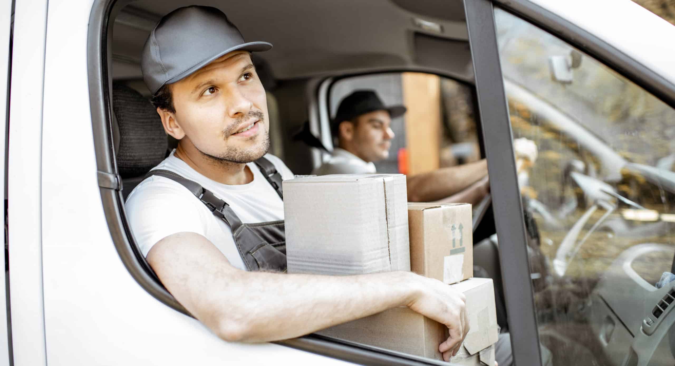 Two delivery men in a van, one is driving, the other is holding parcels and looking out the window
