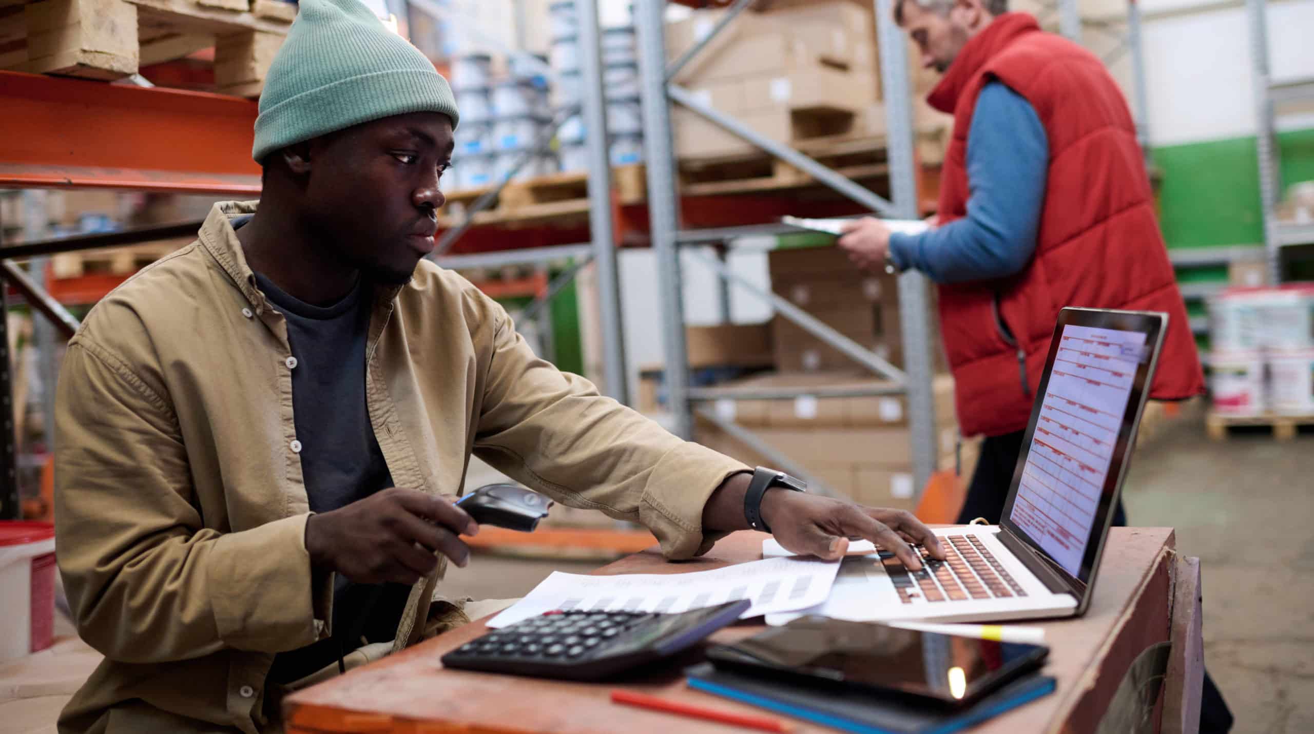 Man in a warehouse using a laptop and also a calculator