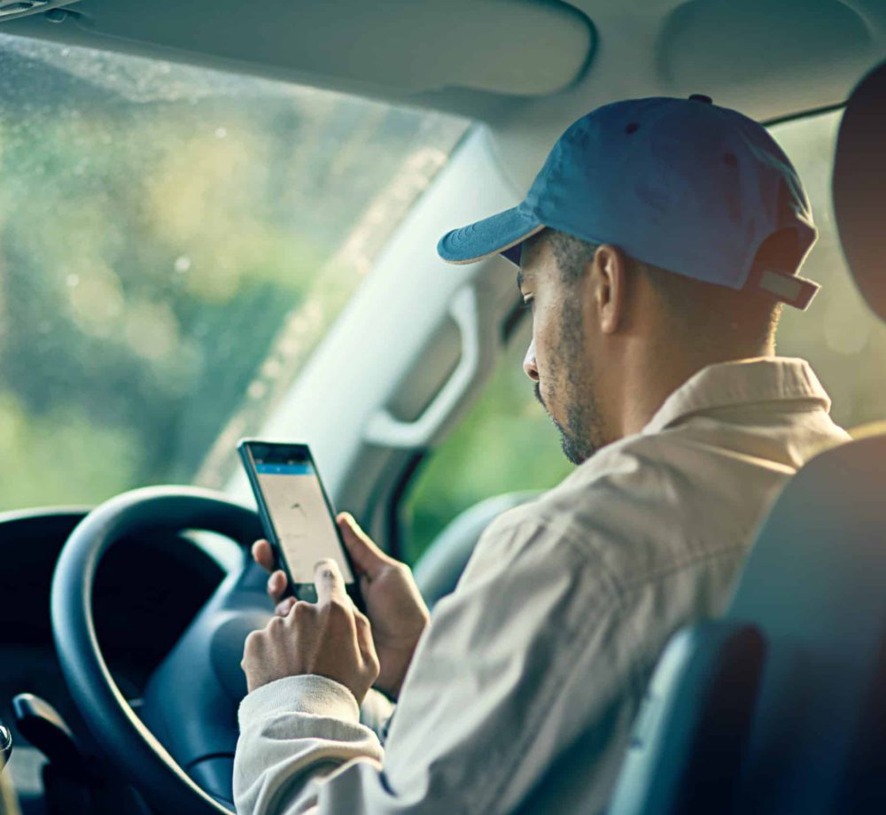 A man wearing a cap uses a smartphone at the wheel of a car