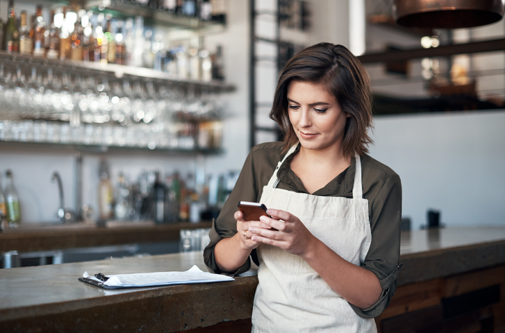 A woman in an apron leans against a bar and looks at her smartphone.