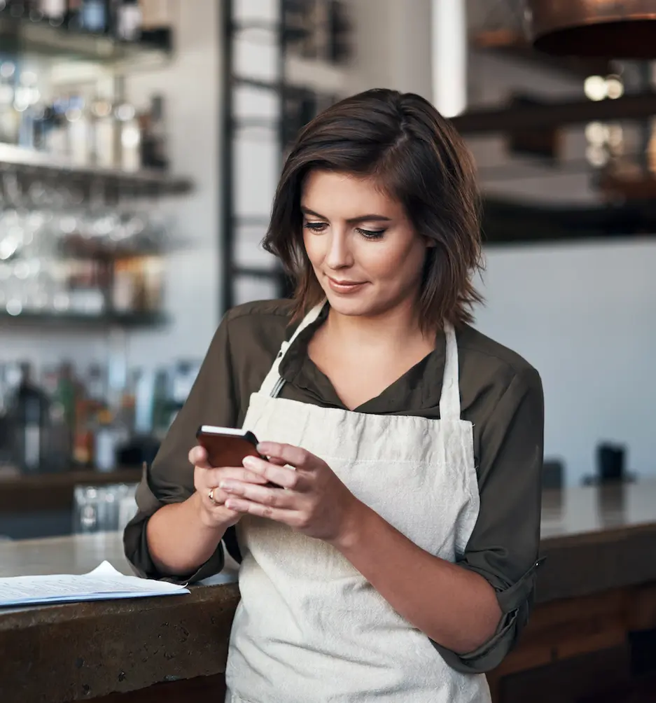 A woman in an apron uses a smartphone while leaning against a bar