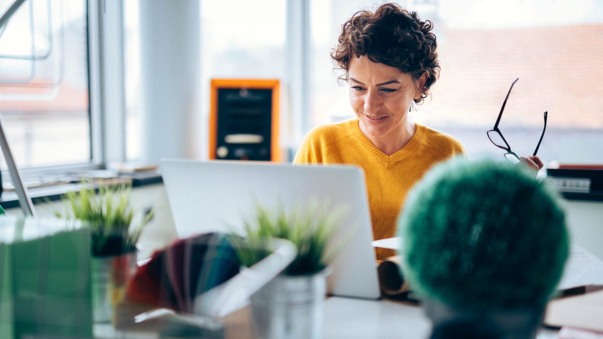 woman sits at desk on her laptop, holding her glasses in her hand