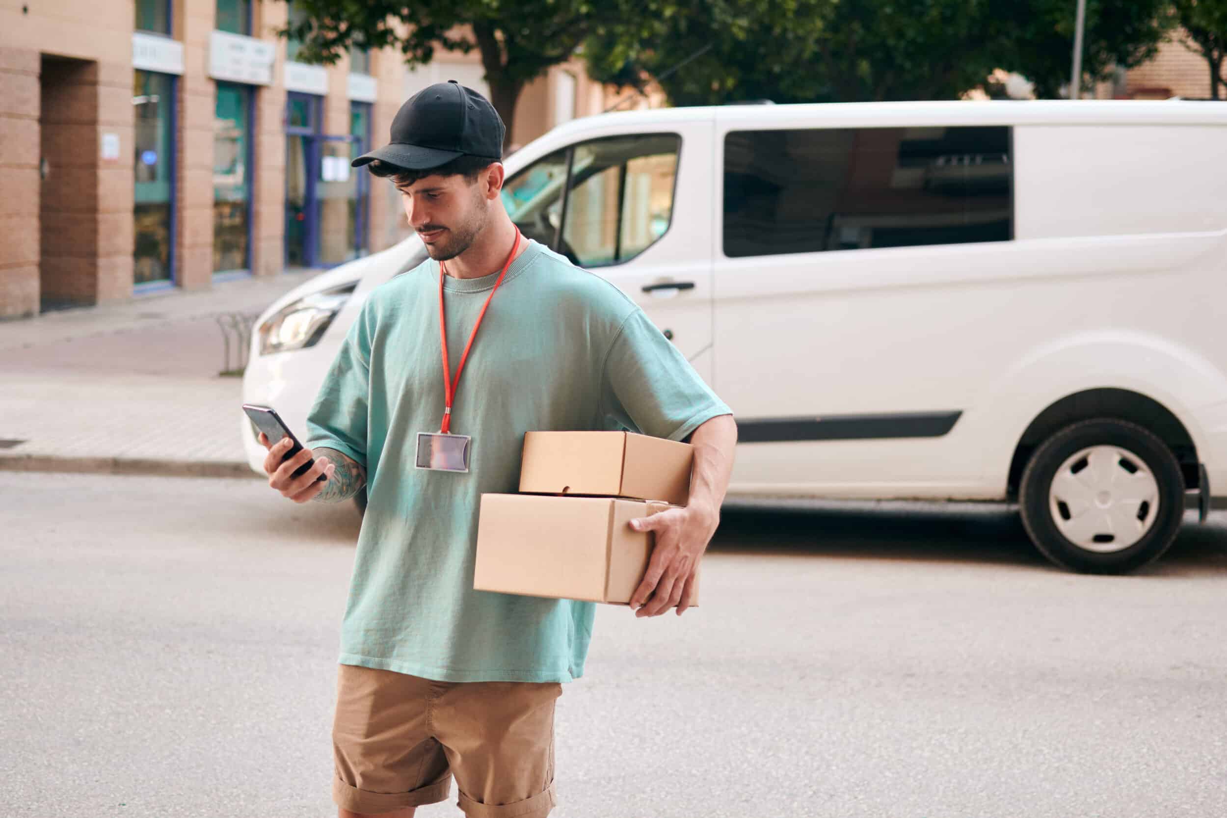 Delivery driver walking infront of his delivery van, holding parcels, and using his mobile device for delivery updates