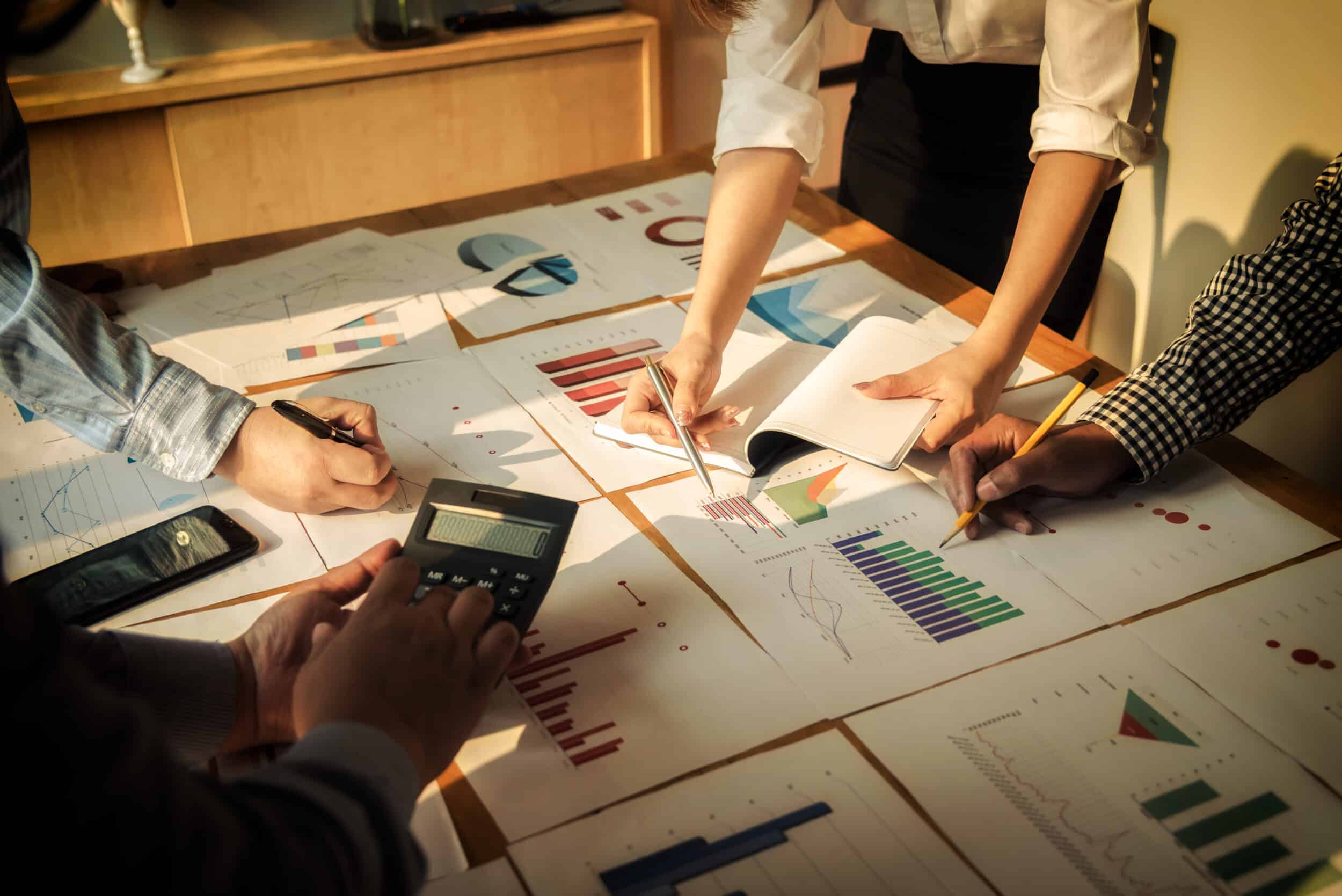 Employees standing around a desk looking at multiple print outs of graphs and pie charts. One person is using a calculator