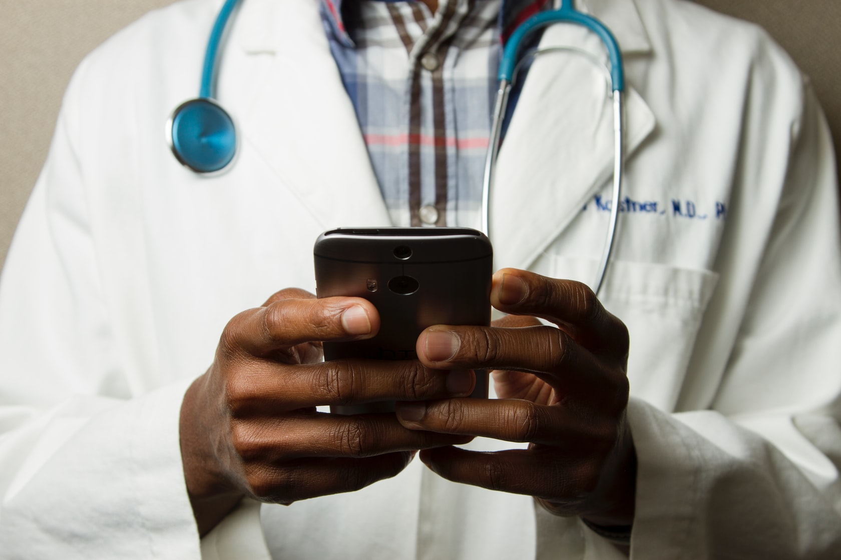 A torso shot of a man wearing a white coat and stethoscope, holding a smartphone.