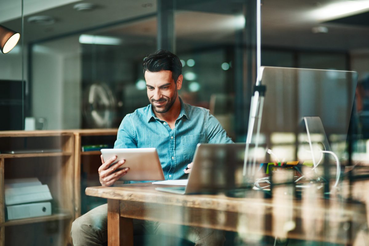 A man sits at a desk and looks at the tablet he is holding