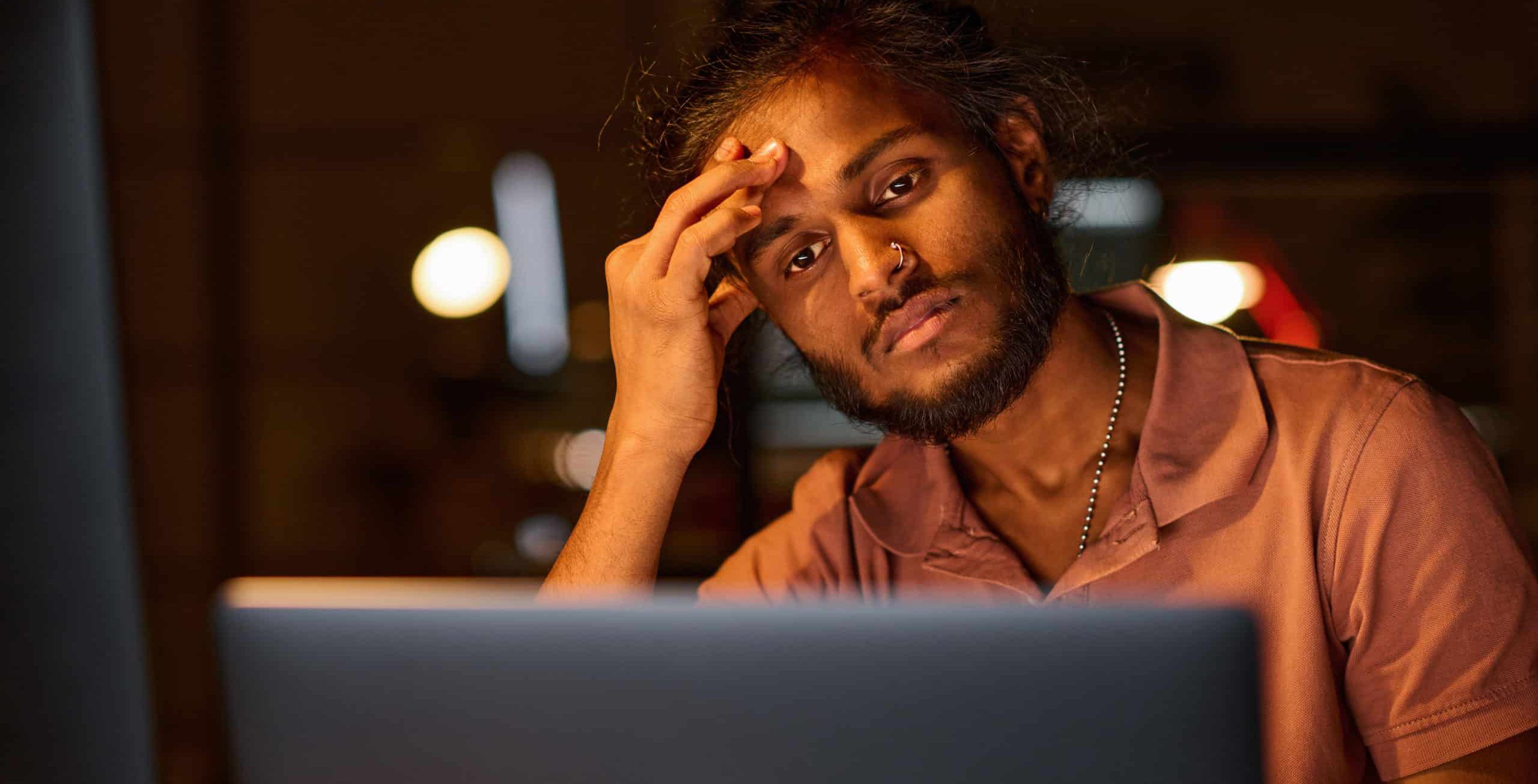 Man sitting infront of a laptop in the evening, looking stressed and tired
